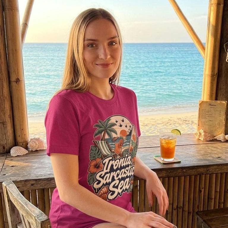Woman sitting at a tropical beach bar wearing the raspberry pink "Ironia, Sarcastic Sea" retro beach T-shirt by SarcasmArt, a funny summer vacation shirt.