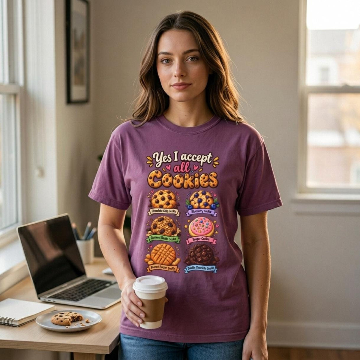 Woman with a coffee at her desk wearing the "Yes, I Accept All Cookies" funny programmer T-shirt by SarcasmArt in Berry – a Comfort Colors 1717 unisex tee with retro script reading Yes, I accept all Cookies above a stack of colorful chocolate chip and frosted cookies.