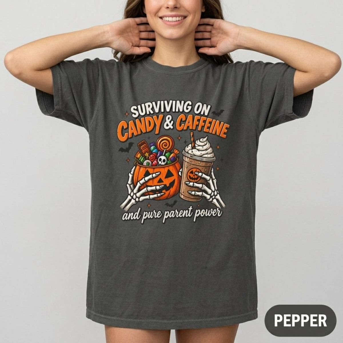 Woman wearing the "Surviving on Candy & Caffeine" Halloween mom T-shirt by SarcasmArt in Pepper (washed charcoal) – a Comfort Colors 1717 unisex tee with a spooky jack-o'-lantern, iced pumpkin coffee and Halloween candy under the words SURVIVING ON CANDY & CAFFEINE.