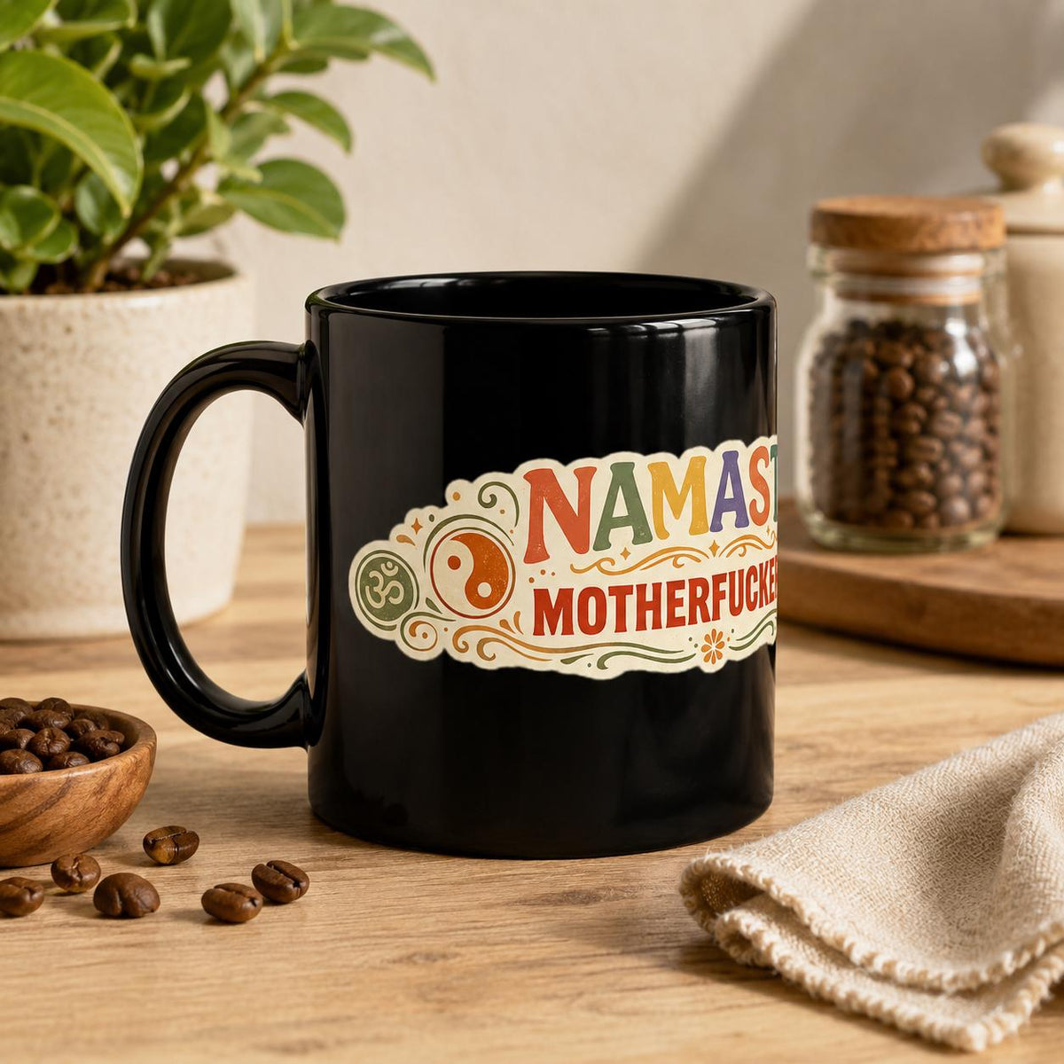"Namaste Motherfuckers" funny yoga coffee mug by SarcasmArt: glossy black ceramic mug with a colorful boho mandala label with the adult-humor phrase NAMASTE MOTHERFUCKERS in red, orange and teal, on a wooden table with coffee beans and a plant.