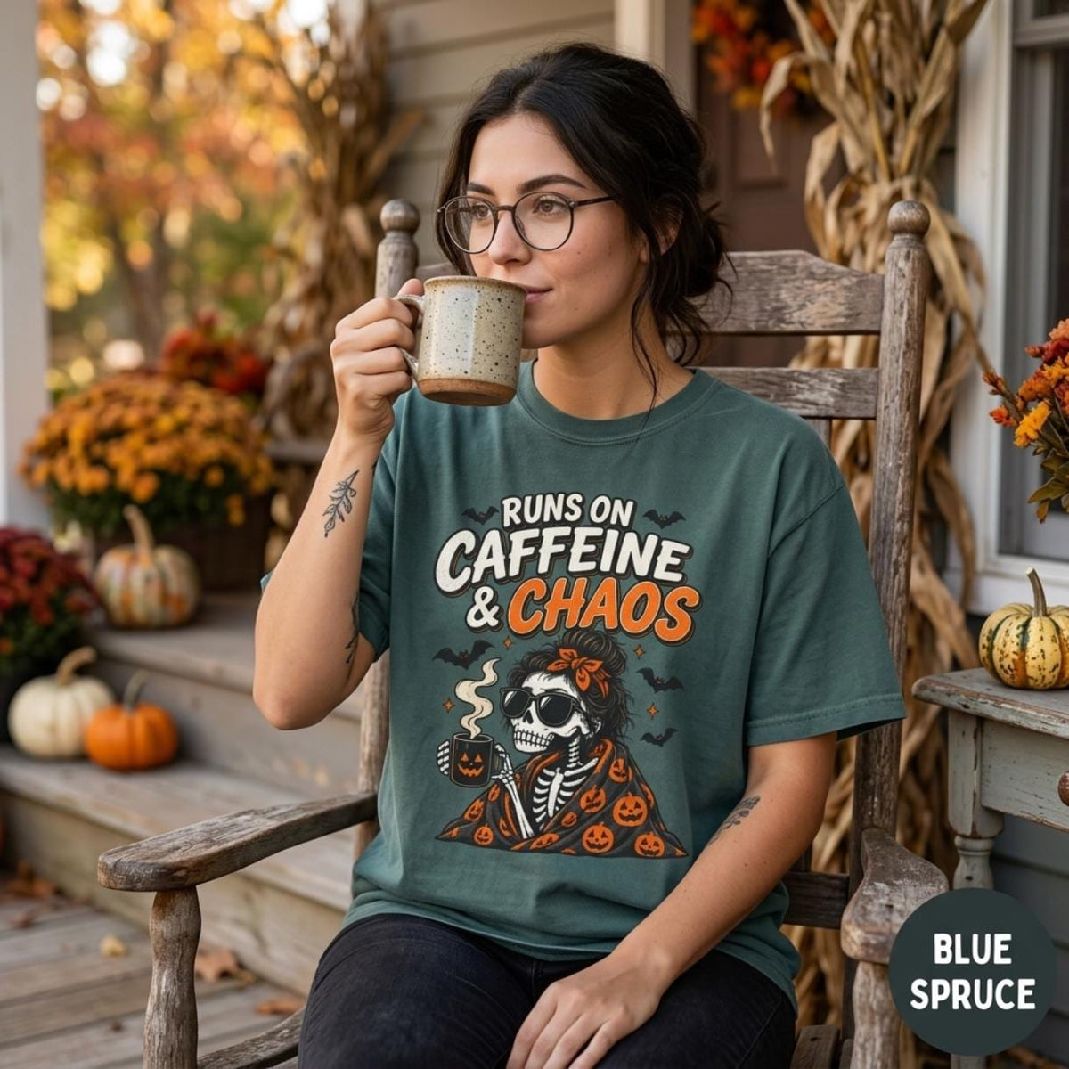 Woman with glasses drinking coffee on a porch with pumpkins, wearing the "Runs on Caffeine & Chaos" skeleton coffee Halloween T-shirt by SarcasmArt in Blue Spruce.