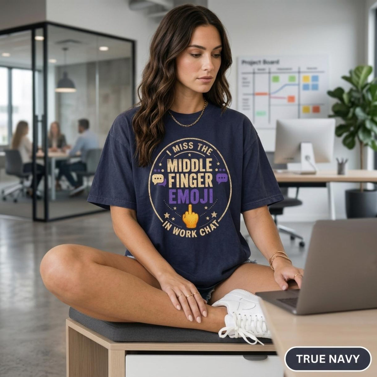 Woman sitting on a desk in a modern office wearing the "I Miss the Middle Finger Emoji in Work Chat" sarcastic office T-shirt by SarcasmArt in True Navy – a funny remote work and coworker gift.