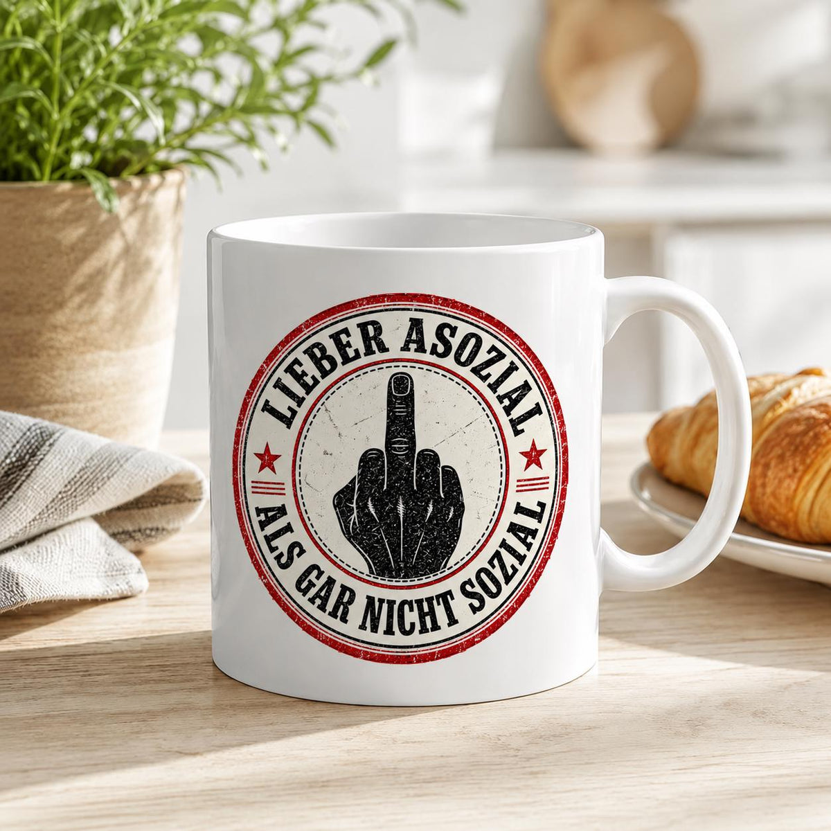 The "Lieber asozial als gar nicht sozial" middle finger coffee mug by SarcasmArt on a breakfast table with a croissant – a funny rude gift with German dark humor.
