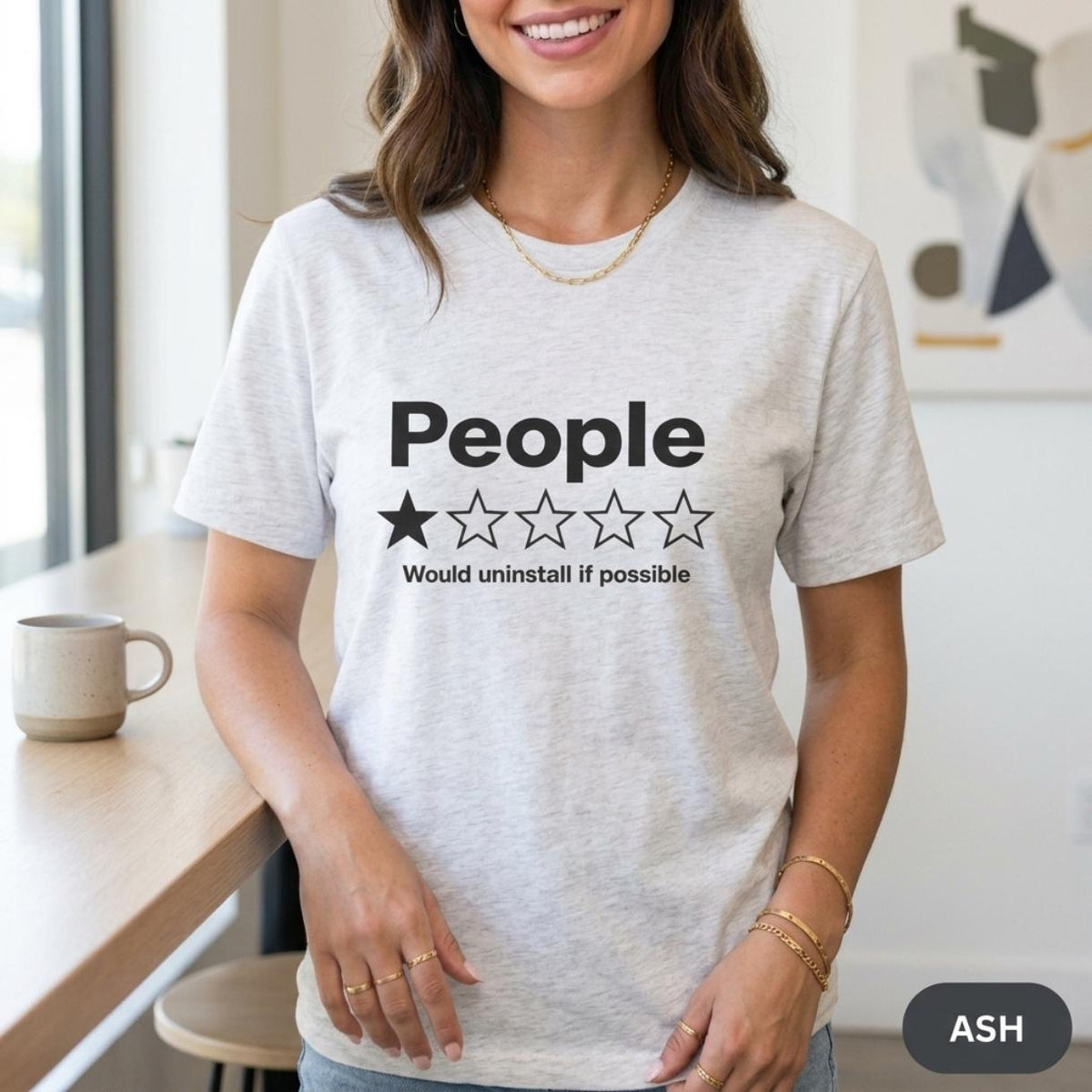 Woman in a bright kitchen wearing the "People – 1 Star, Would Uninstall If Possible" funny review T-shirt by SarcasmArt in Ash (light heather grey).