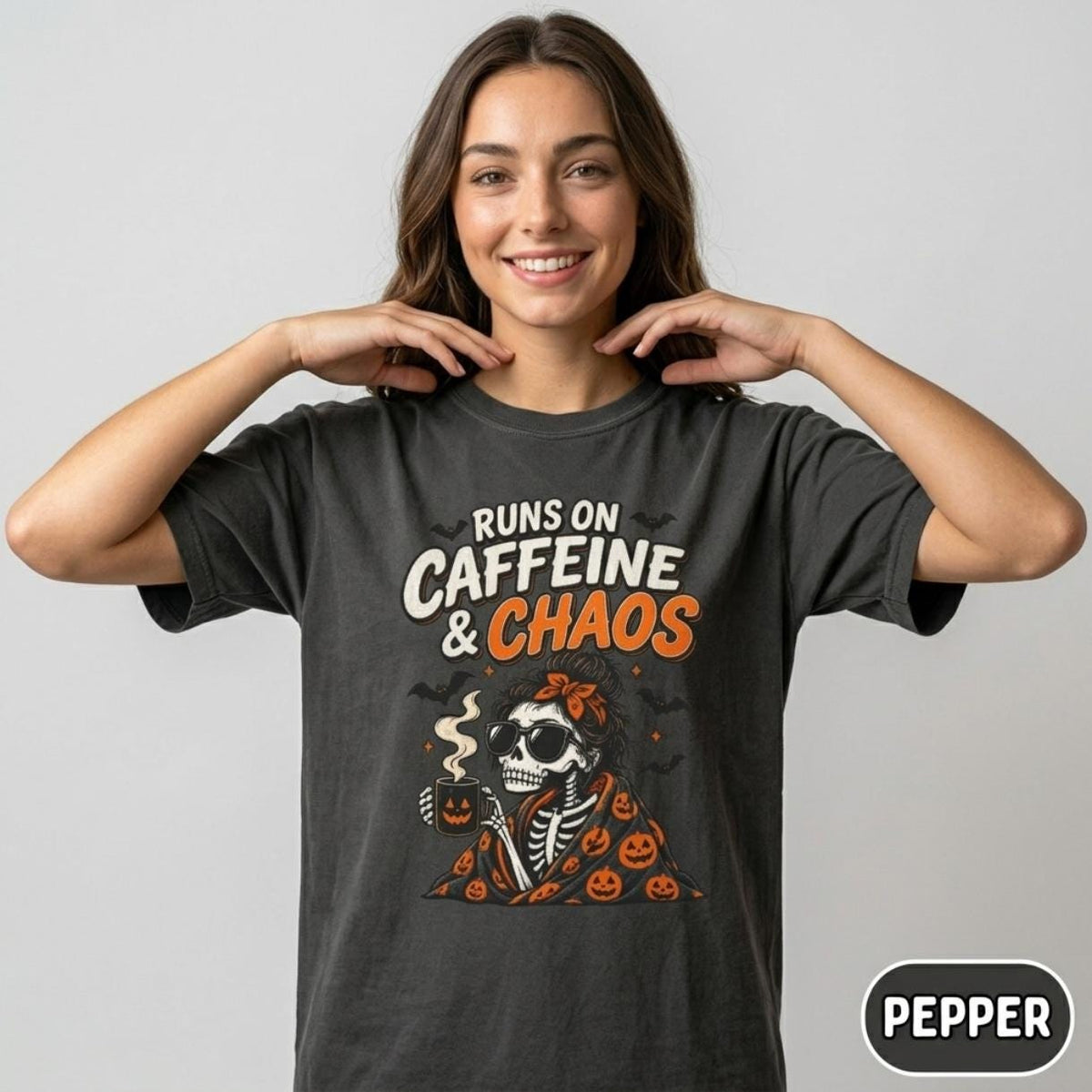 Smiling woman wearing the "Runs on Caffeine & Chaos" skeleton coffee Halloween T-shirt by SarcasmArt in Pepper (washed charcoal) – a Comfort Colors 1717 unisex tee with a grinning skeleton sipping coffee over pumpkins with RUNS ON CAFFEINE & CHAOS lettering in cream and orange.