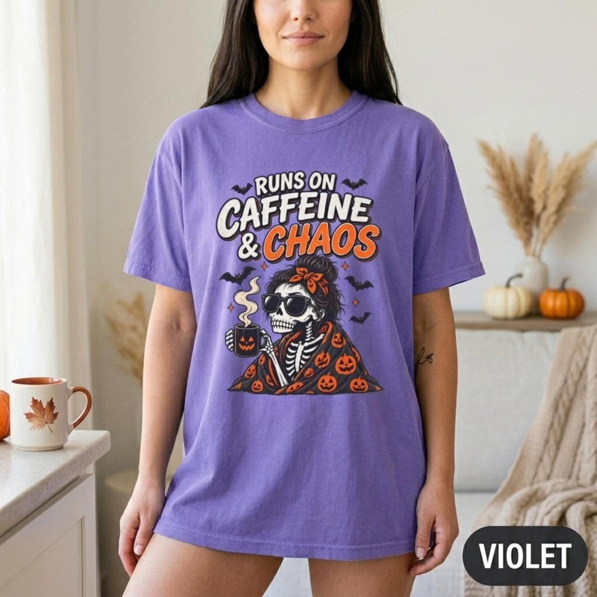 Woman in a fall kitchen wearing the "Runs on Caffeine & Chaos" skeleton coffee Halloween T-shirt by SarcasmArt in Violet as an oversized tee.