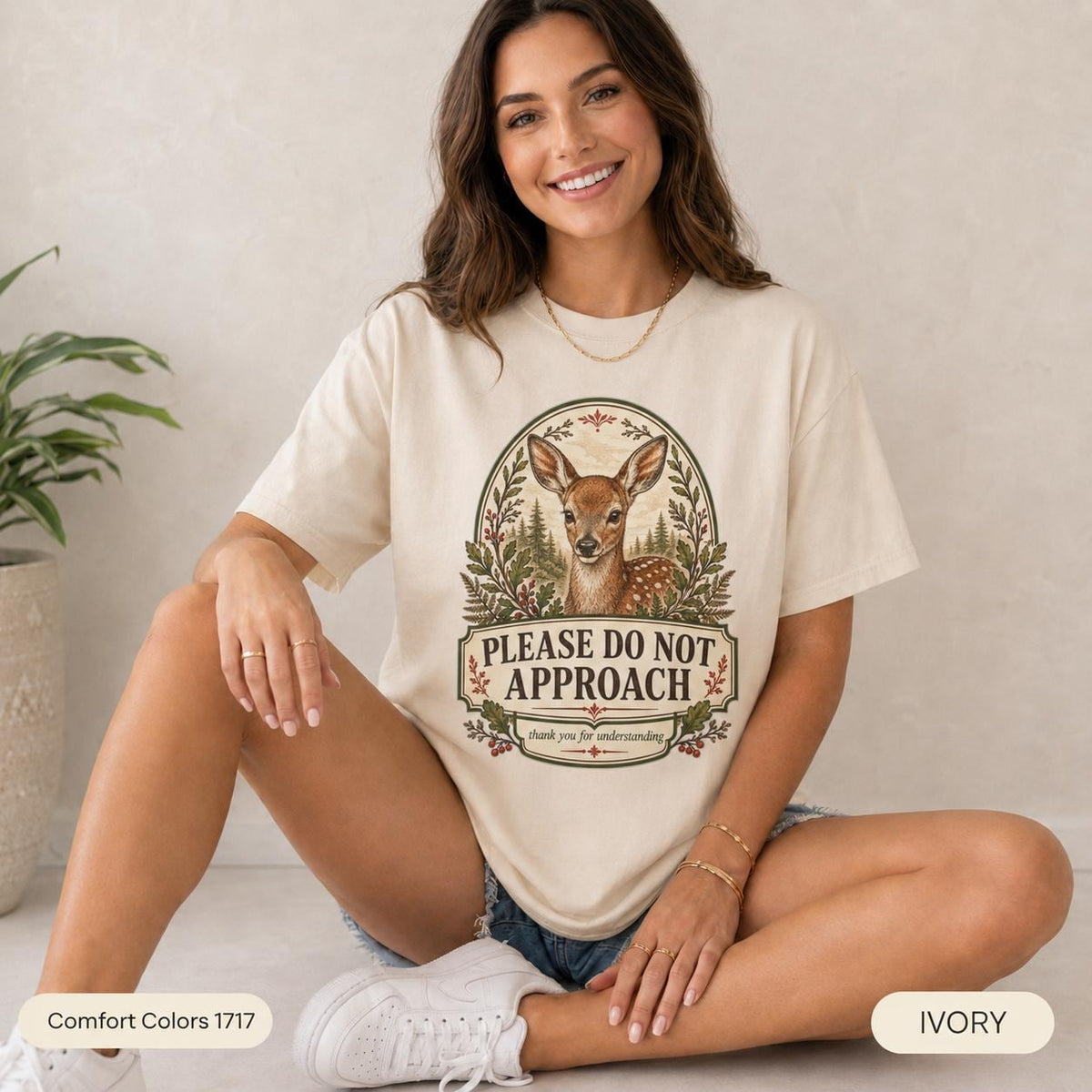 Smiling woman sitting on the floor wearing the "Please Do Not Approach" vintage fawn introvert T-shirt by SarcasmArt in Ivory – a relaxed unisex forestcore tee with a vintage woodland deer fawn in an oval frame of ferns and wildflowers above the words PLEASE DO NOT APPROACH.
