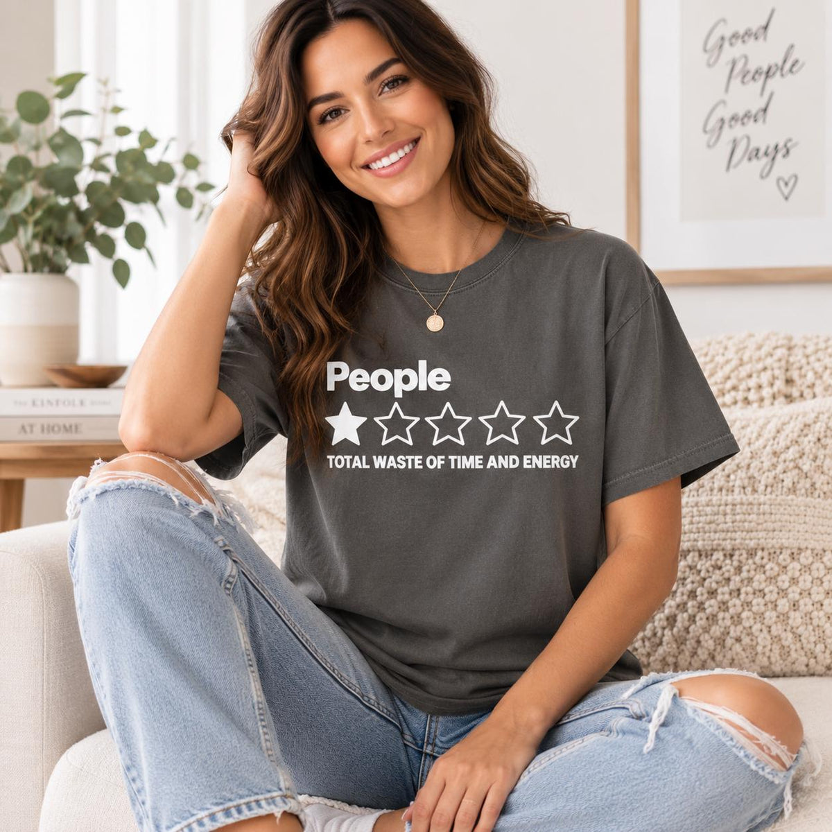 Smiling woman sitting on a sofa wearing the "People – 1 Star, Total Waste of Time and Energy" sarcastic review T-shirt by SarcasmArt in Pepper (washed charcoal) – a funny introvert gift.