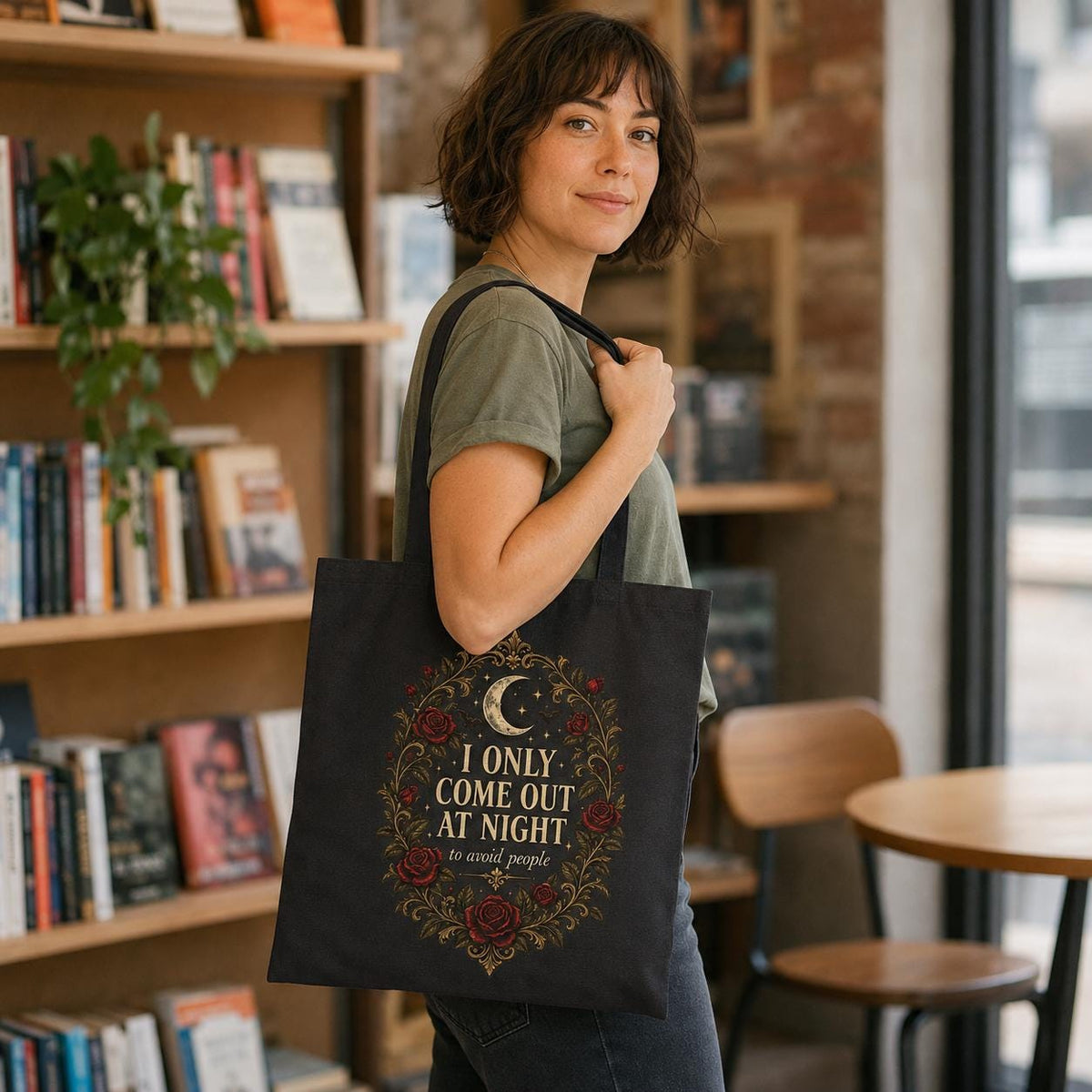 Woman in a bookstore café carrying the Black "I Only Come Out at Night to Avoid People" gothic canvas tote bag by SarcasmArt.