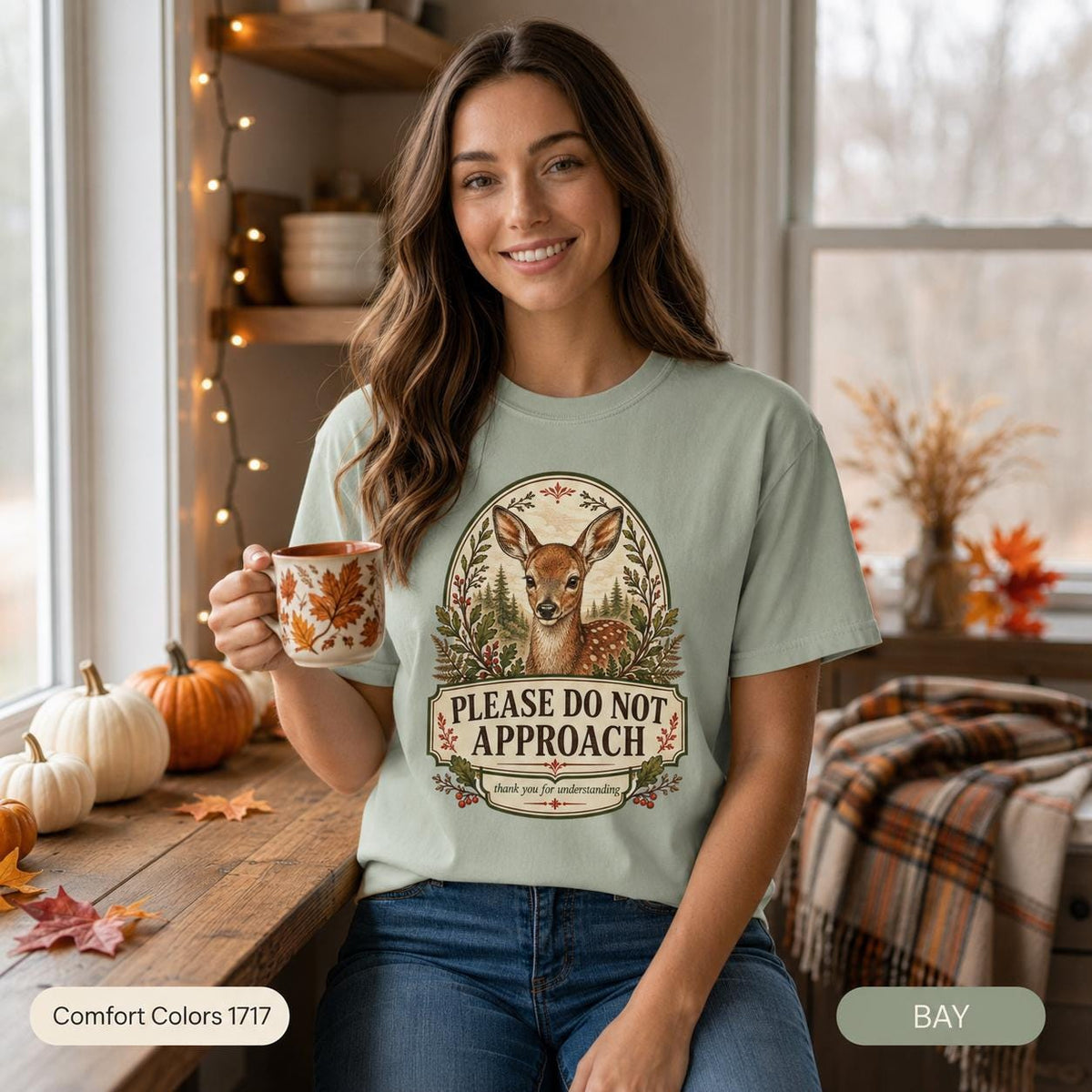 Woman holding a cup of tea in a cozy autumn kitchen wearing the "Please Do Not Approach" vintage fawn introvert T-shirt by SarcasmArt in Bay (sage green) – a funny homebody gift.