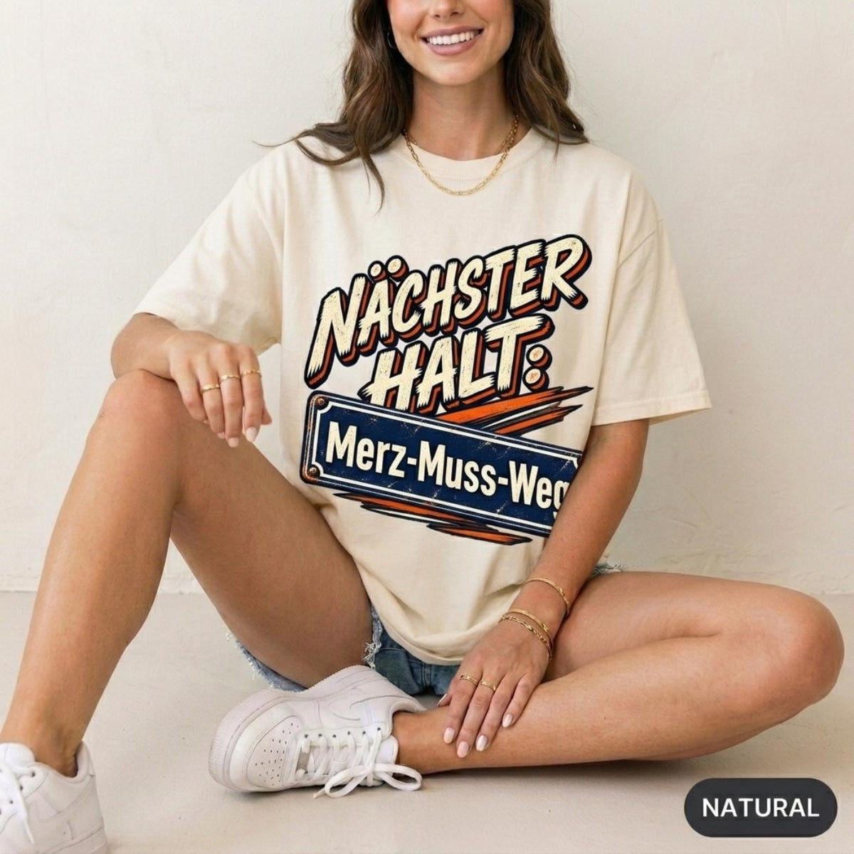 Woman sitting on the floor wearing the "Nächster Halt: Merz-Muss-Weg" German political satire T-shirt by SarcasmArt in Natural – a Gildan Softstyle 64000 tee with retro comic lettering NÄCHSTER HALT: above a blue street-sign style plate reading Merz-Muss-Weg with orange speed lines.