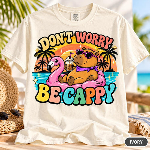 Ivory "Don't Worry Be Cappy" capybara beach T-shirt by SarcasmArt on a beach lounge chair – Comfort Colors 1717 unisex tee with a relaxed capybara in sunglasses floating on a pink flamingo ring with a cocktail, palm trees and a retro sunset, with colorful DON'T WORRY BE CAPPY lettering.