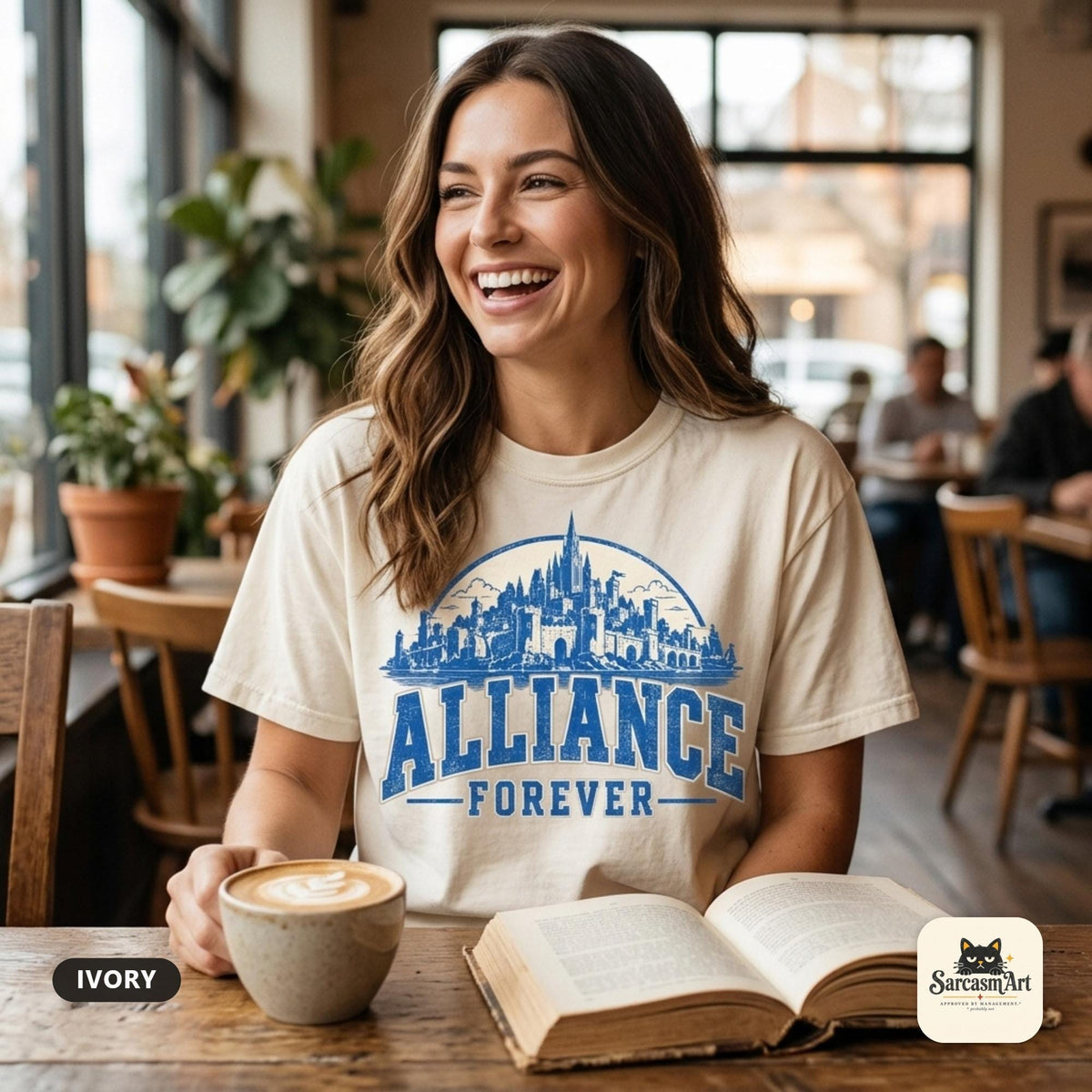 Laughing woman with a latte and an open book in a café wearing the "Alliance Forever" World of Warcraft gamer T-shirt in Ivory – a Comfort Colors 1717 unisex tee by SarcasmArt with blue distressed ALLIANCE FOREVER varsity lettering under a fantasy city skyline.