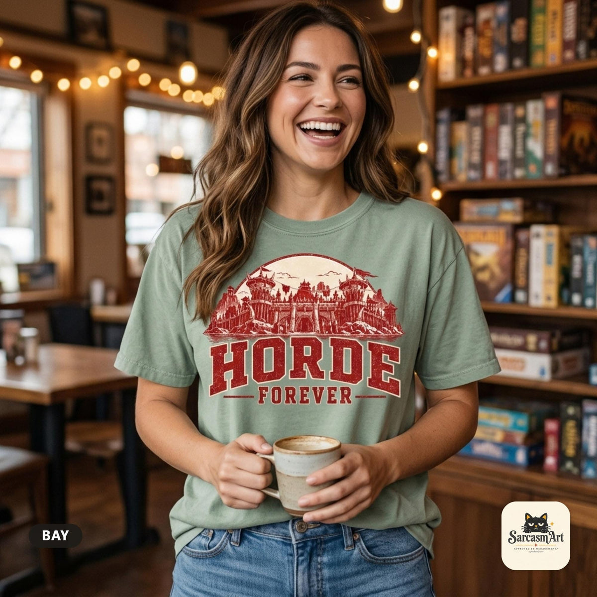 Smiling woman holding a coffee mug in a cozy board game café wearing the "Horde Forever" World of Warcraft gamer T-shirt in Bay (sage green) – a Comfort Colors 1717 unisex tee by SarcasmArt with red distressed HORDE FOREVER varsity lettering over a gothic fantasy castle skyline.