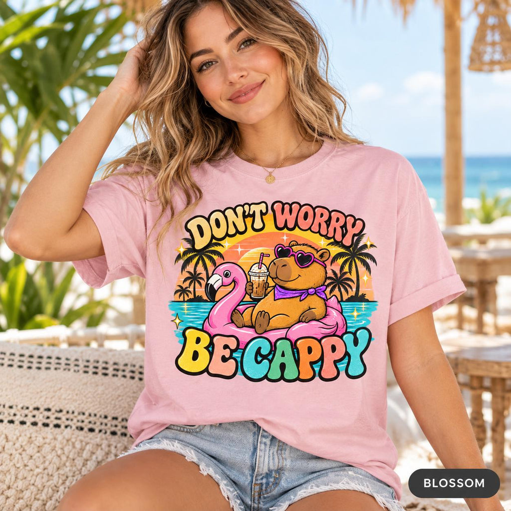 Woman in sunglasses at a beach bar wearing the "Don't Worry Be Cappy" capybara beach T-shirt by SarcasmArt in Blossom (soft pink).