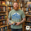 Bearded man holding a board game in a game store wearing the "Alliance Forever" World of Warcraft gamer T-shirt in Bay (sage green) – a Comfort Colors 1717 unisex tee by SarcasmArt with blue distressed ALLIANCE FOREVER varsity lettering under a fantasy city skyline.