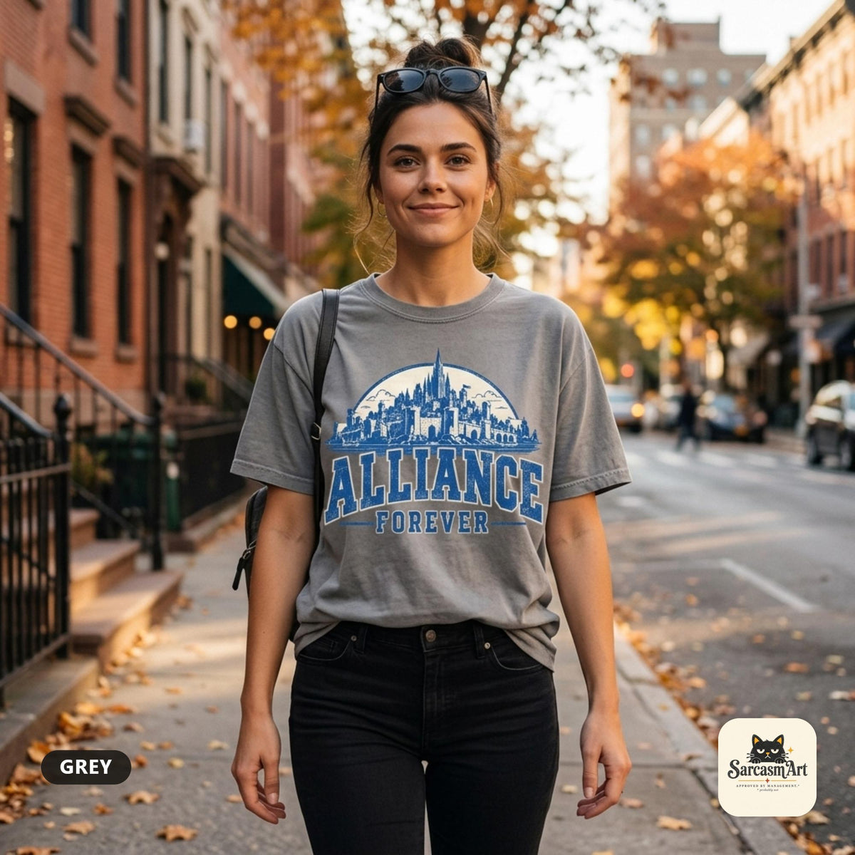 Woman walking down an autumn city street wearing the "Alliance Forever" World of Warcraft gamer T-shirt in Grey – a Comfort Colors 1717 unisex tee by SarcasmArt with blue distressed ALLIANCE FOREVER varsity lettering under a fantasy city skyline.