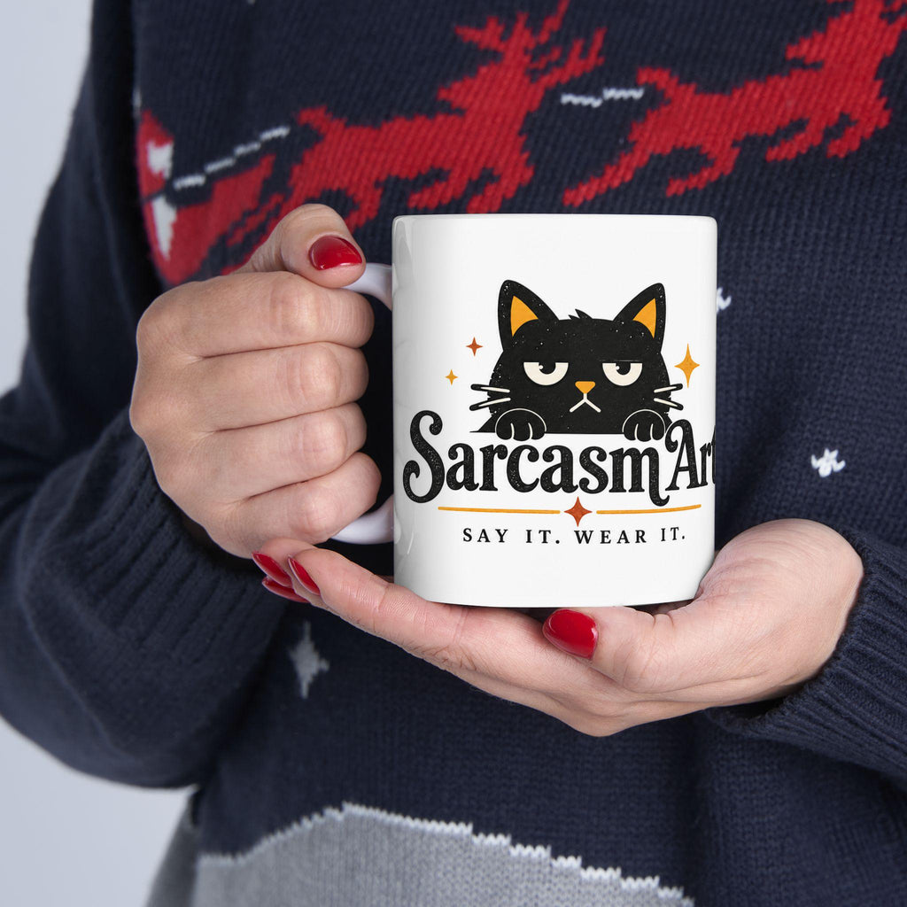 May include: A white ceramic mug featuring a graphic of a black cat with yellow inner ears and a grumpy expression. Below the cat, the text reads SarcasmArt in a stylized black font, followed by the smaller text SAY IT. WEAR IT. in black.